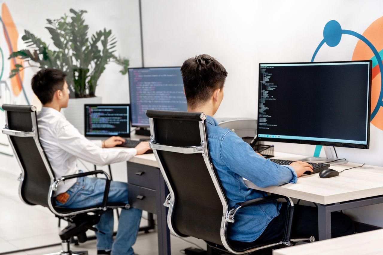 heros-img Two male developers at desks programming in a modern office workspace with large monitors.